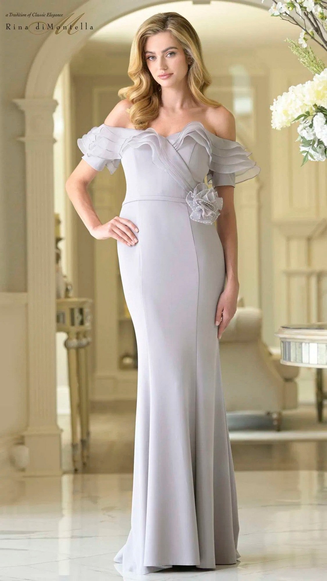 A woman stands indoors wearing the Rina Di Montella RD3418, a grey floor-length formal dress with an off-the-shoulder neckline, ruffled sleeves, and a floral accent at the waist.
